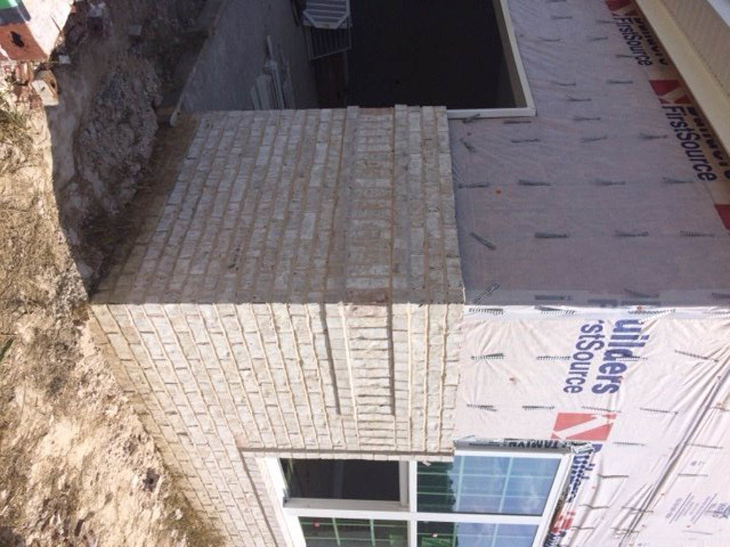 Red brick house under construction with a window, plastic sheeting covering part of the wall, and a white tarp displaying blue and red text.