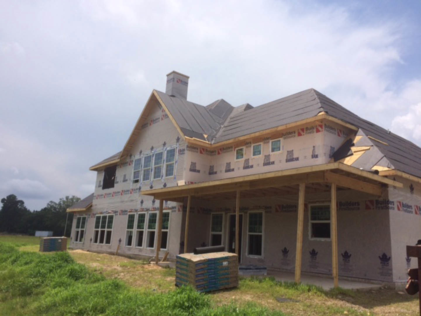 Two-story home under construction with exposed framing, spacious covered porch, white-framed windows, chimney, stacks of pallets and cardboard boxes on grassy yard, cloudy sky