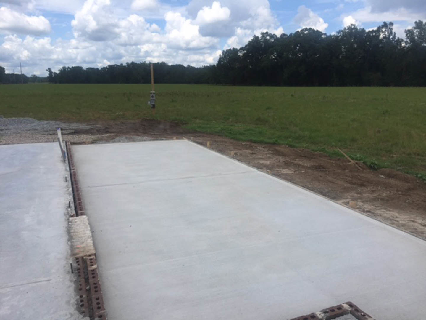 Concrete walkway bordered by grass and dirt, drain cover visible in foreground, cloudy sky above trees in background