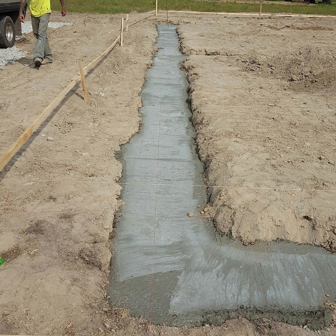 Man in yellow shirt and grey pants standing beside dirt trench on grassy outdoor home site, tire visible in foreground