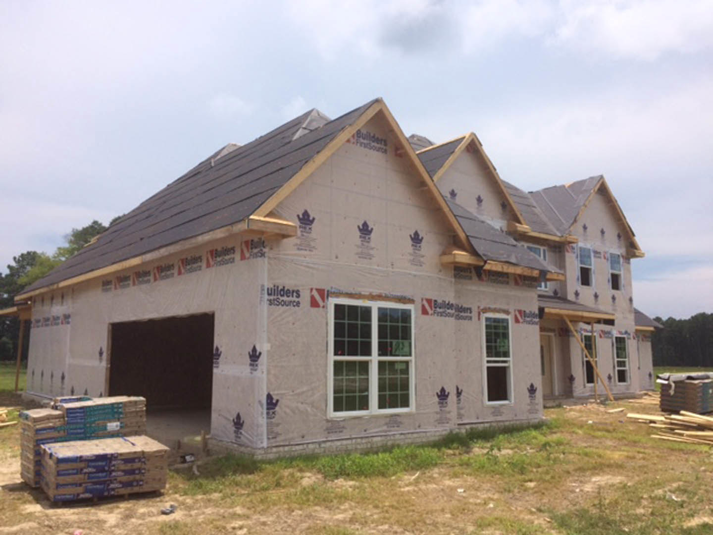 Partially built house with exposed framing, attached garage, white-framed windows, construction materials stacked on pallets, cloudy sky overhead