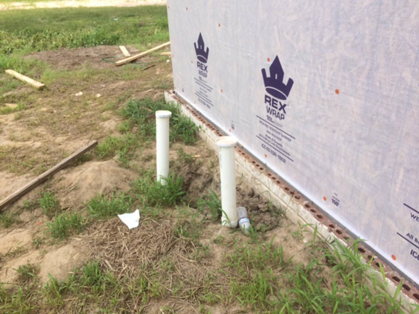White plastic pipes lying next to a white wall with blue and purple logos, a white paper on grass, wooden beam on ground, white pole with green leaf, and purple crown logo on white