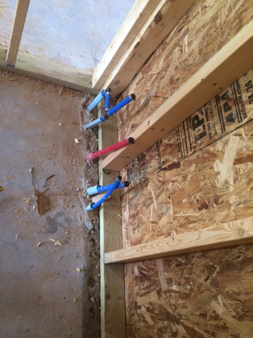 Exposed wood framing with blue plumbing pipes running along beams and walls, red object partially visible, unfinished dirt floor, construction materials and insulation in