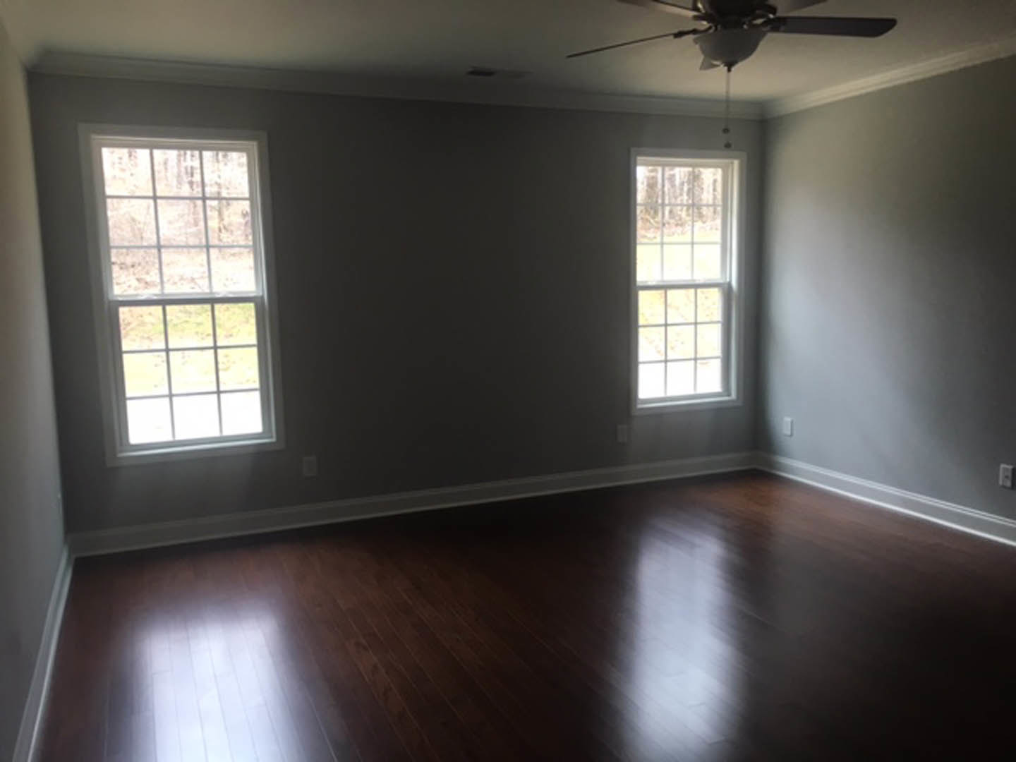 Dark wood floor with white baseboards, multi-pane window, ceiling fan with light fixture, upholstered chair in corner, neutral wall color.