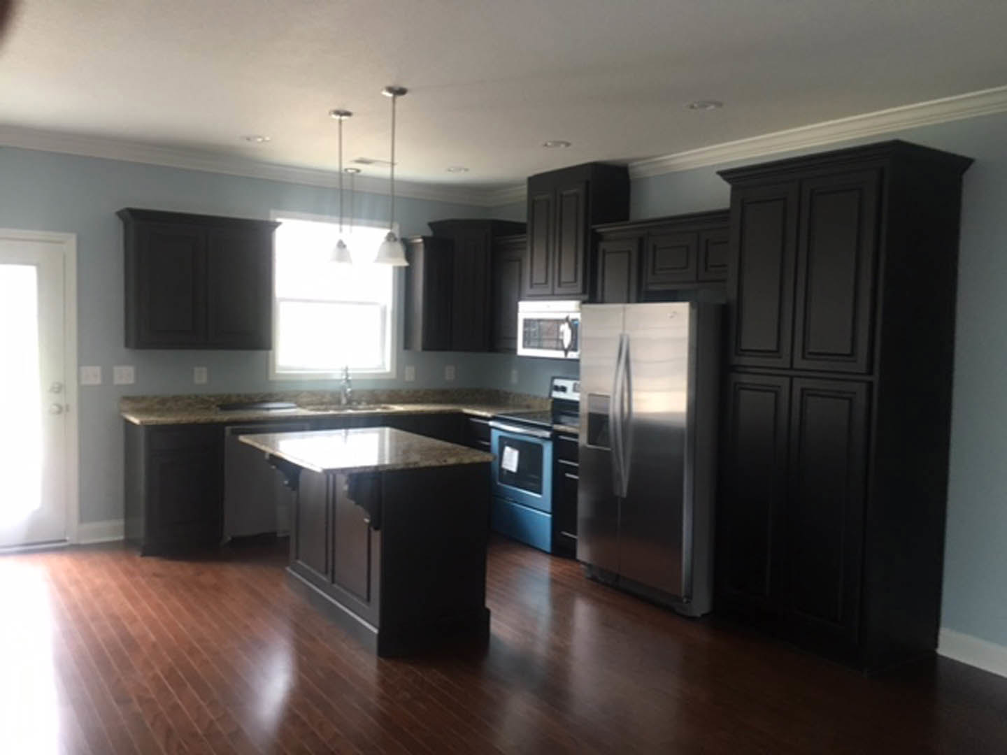 Black kitchen cabinets, stainless steel refrigerator, marble-topped island, blue oven, light-colored flooring, and white walls