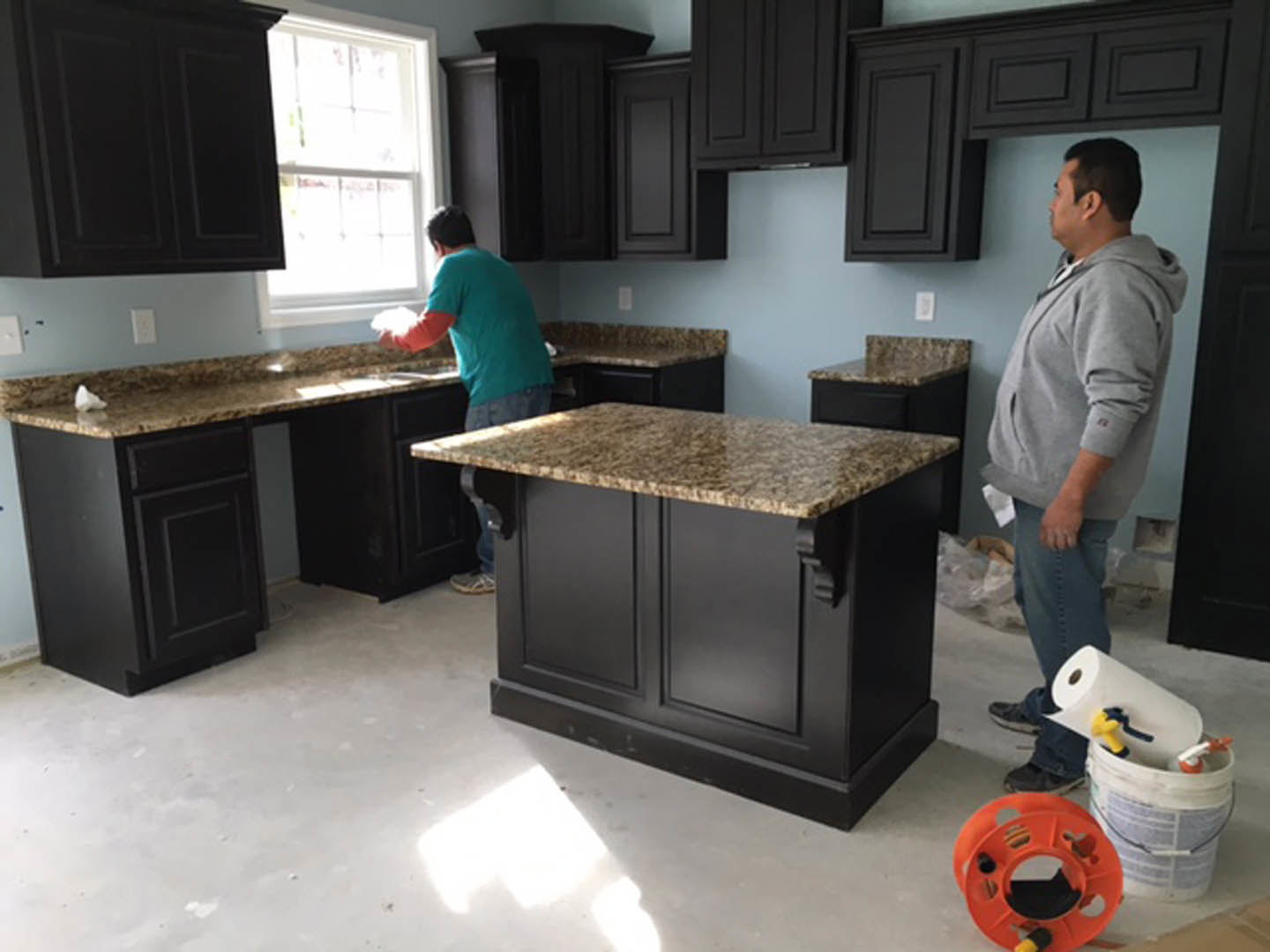 Two men standing in a modern kitchen with white cabinetry, black marble countertop, stainless steel appliances, and a paint bucket with spray gun on the island.