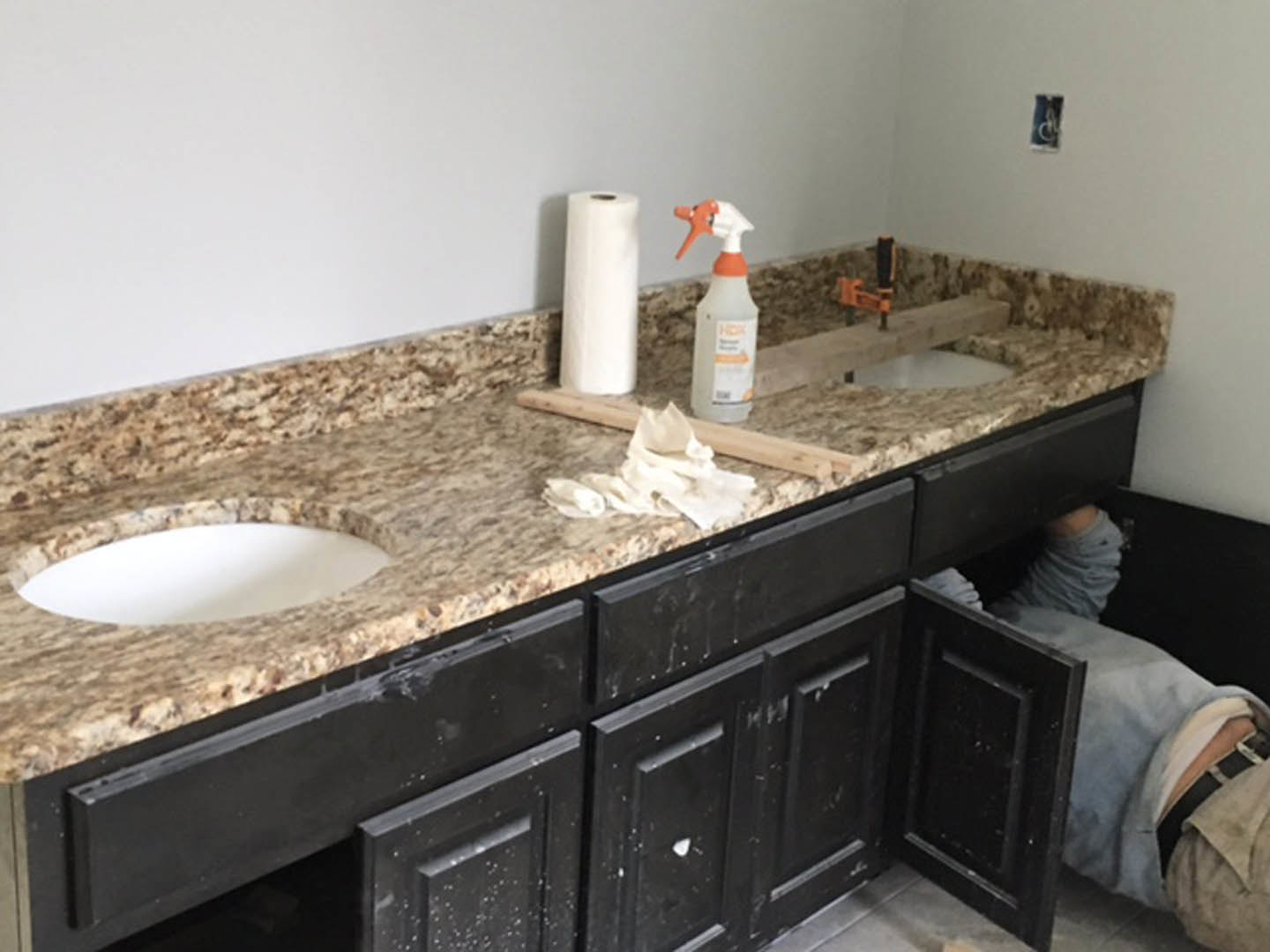 Person crouched beneath white kitchen sink cabinet, hand reaching toward plumbing fixtures, orange-capped bottle and paper towel roll visible on tiled floor, white cabinetry and