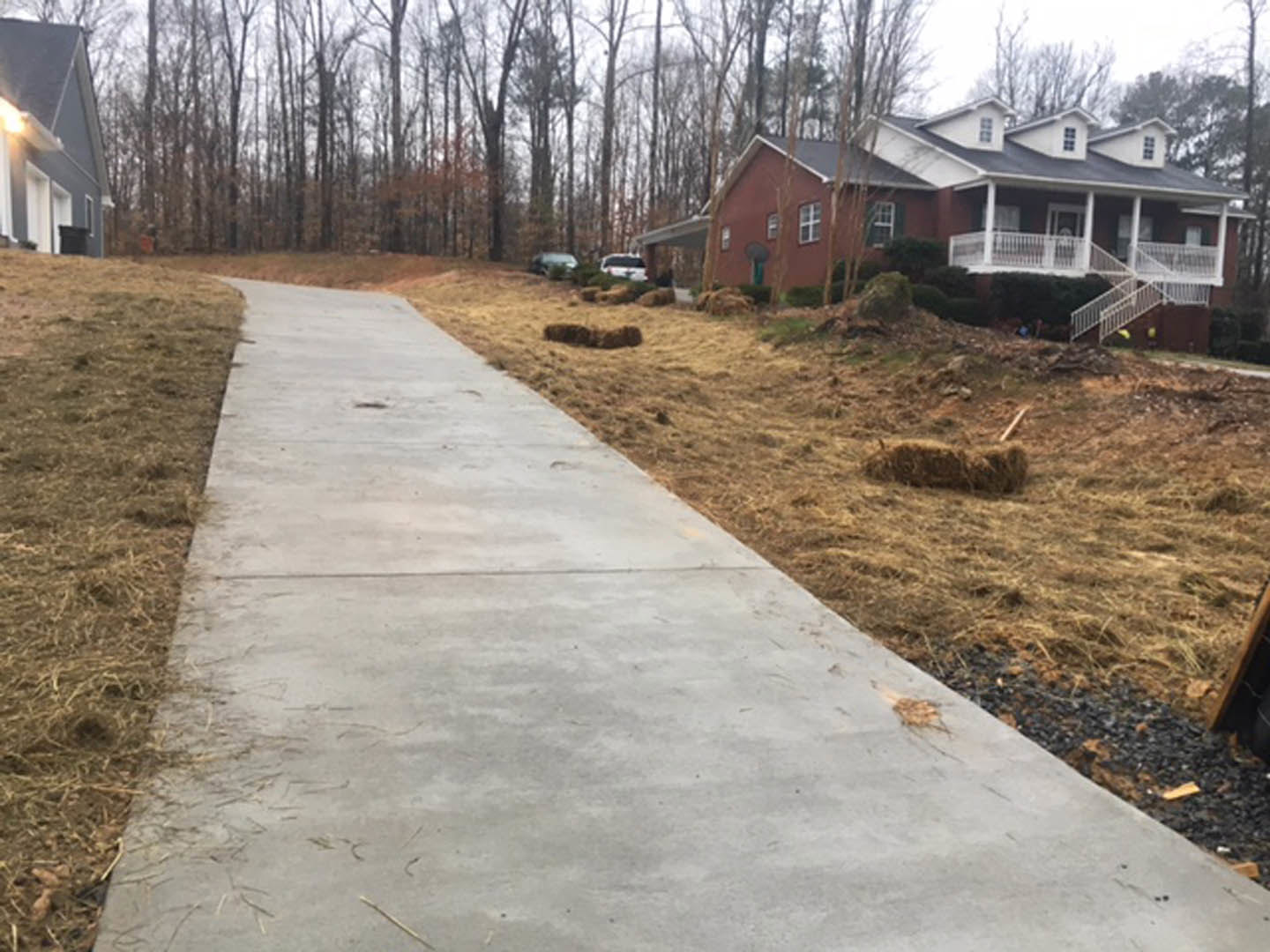 Concrete walkway bordered by green grass leading to a house with white railings, trees in the background, and a clear sky overhead