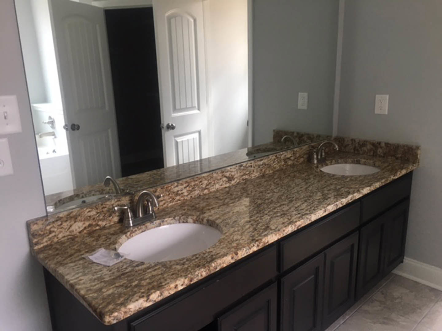 Bathroom with double vanity featuring undermount sinks, quartz countertop, large frameless mirror, chrome faucets, white tile backsplash, and visible electrical outlet on wall