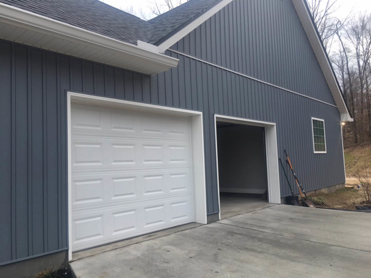 White paneled garage door set in gray siding, concrete driveway in foreground, white-framed window adjacent to door