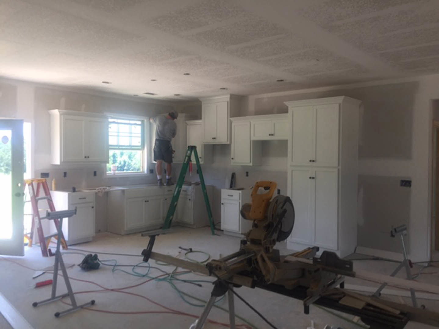 Man on a ladder installing or repairing near white kitchen cabinets and window, with plaster walls and ceiling in a residential renovation setting