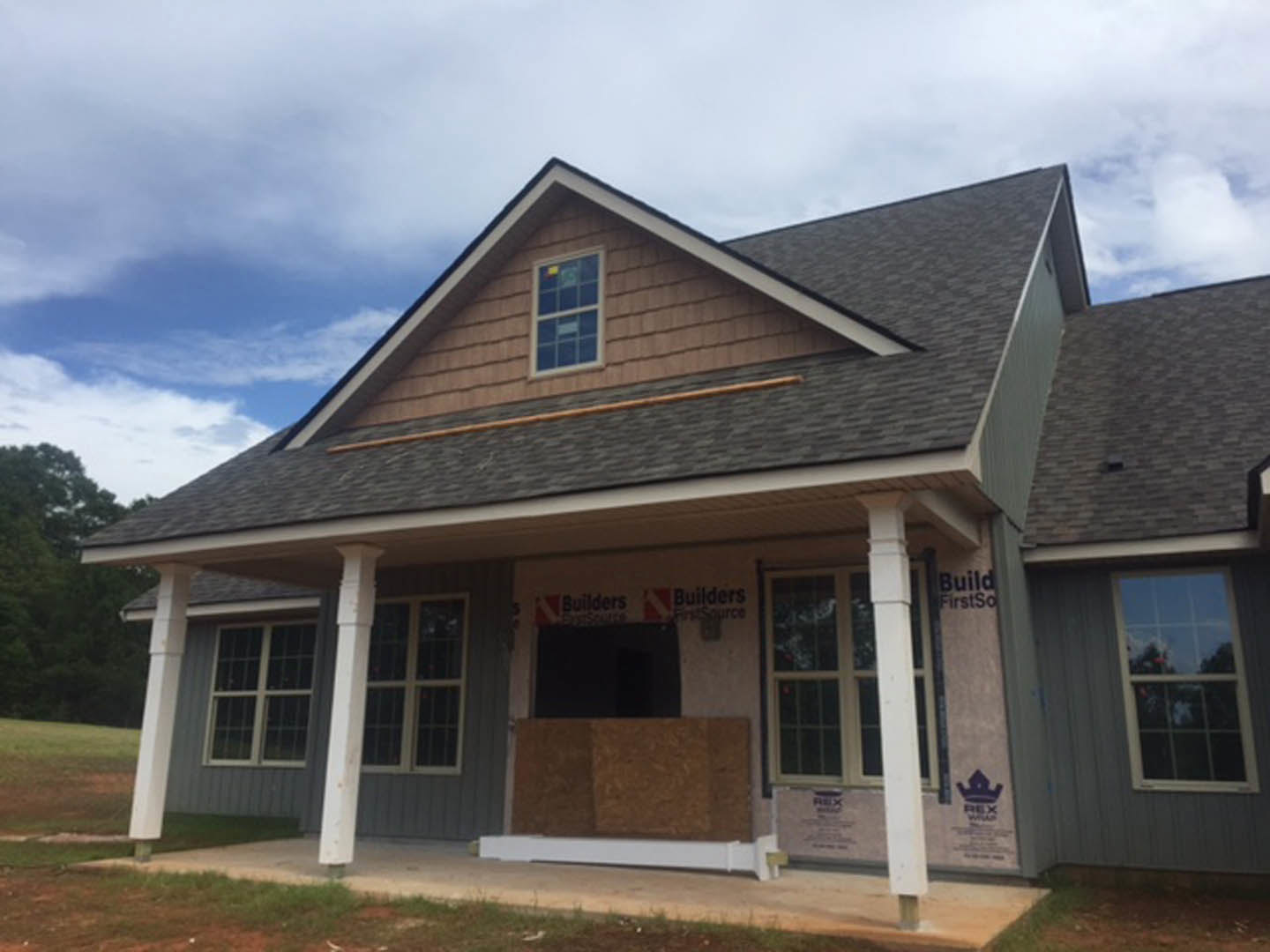 Framed house under construction with exposed wooden siding, white window frames, partially finished roof, and clear blue sky overhead