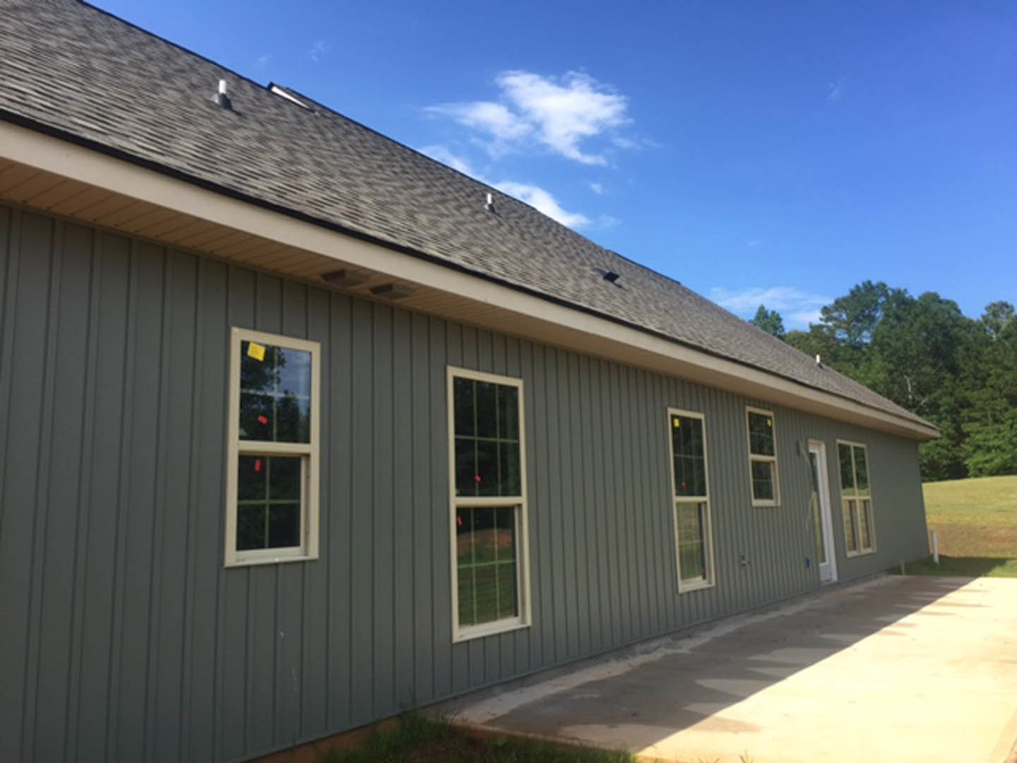 Two-story house with light siding, multiple windows, shingled roof, paved driveway, and trees in the background under a clear blue sky
