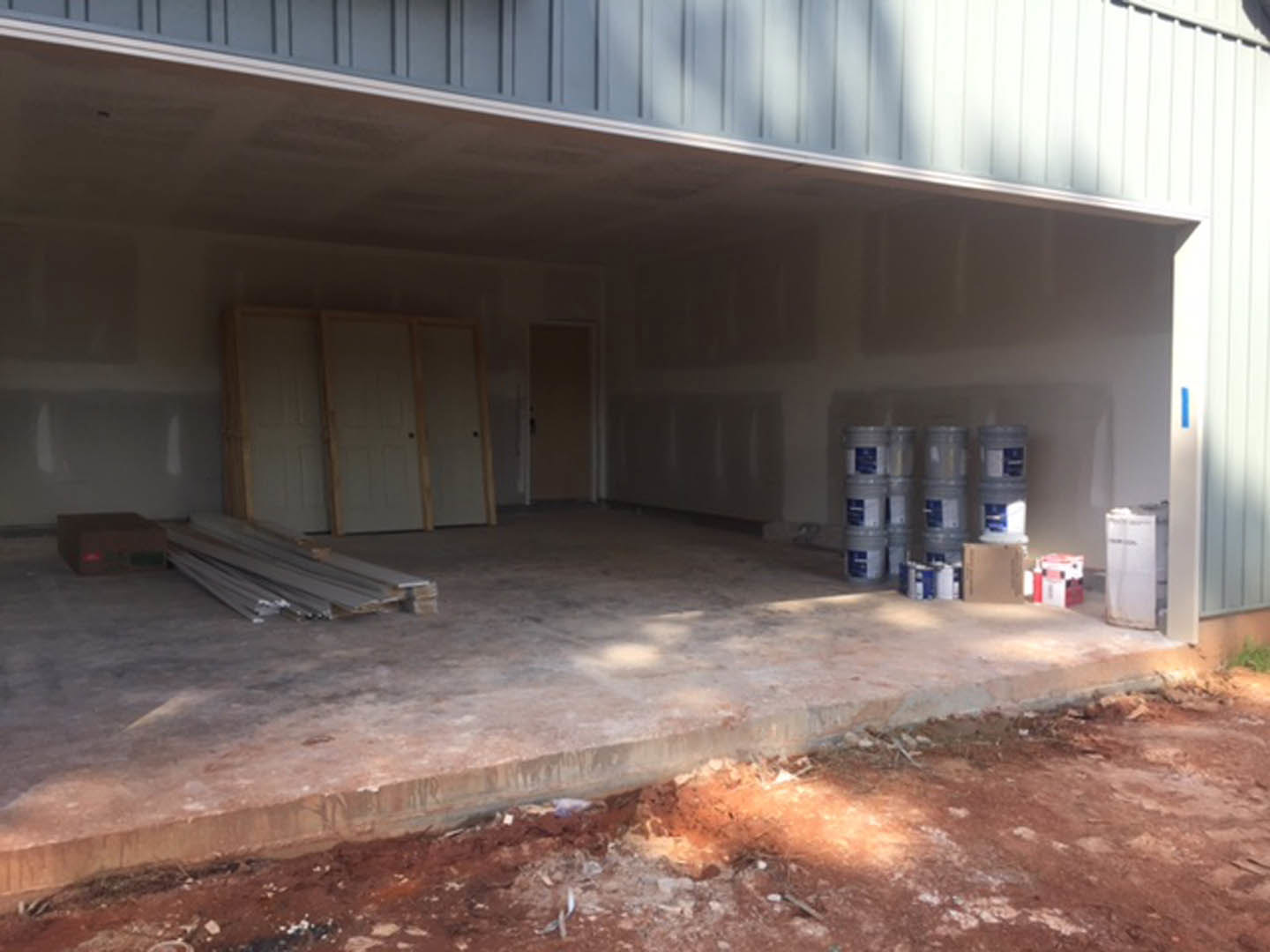 Garage interior with numerous wooden barrels, stacked metal bars, white doors along one wall, concrete slab flooring, and several paint buckets.