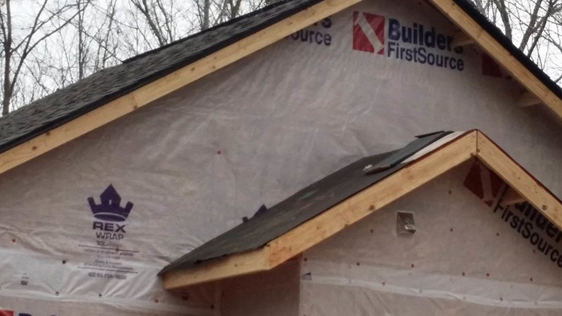 Partially built house with exposed framing, plastic sheeting covering roof, white siding, and surrounding leafless trees under a cloudy sky