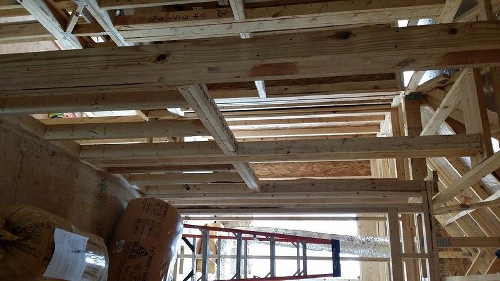 Wooden ceiling beams and metal framing in a partially constructed home interior with a ladder, insulation materials, and construction supplies scattered on the floor.