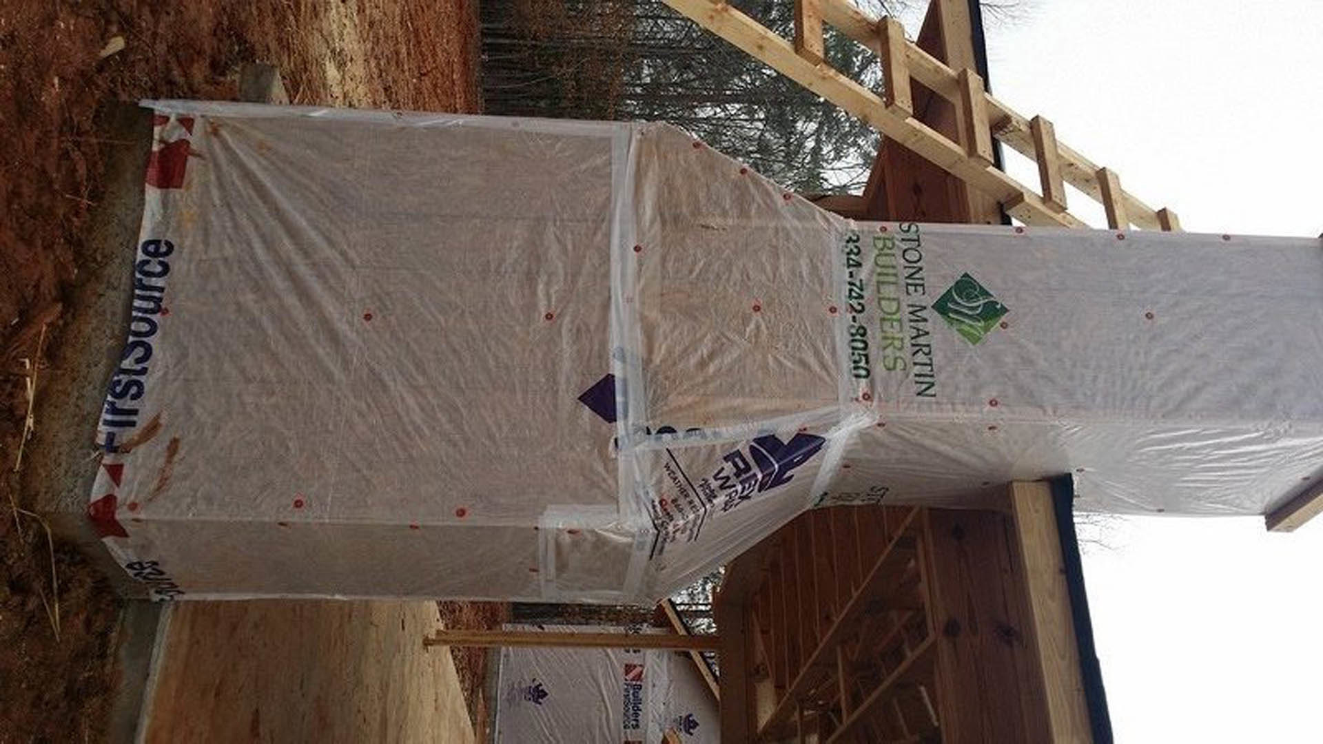 Partially constructed house wrapped in white plastic sheeting, exposed framing visible beneath, set outdoors under a clear sky