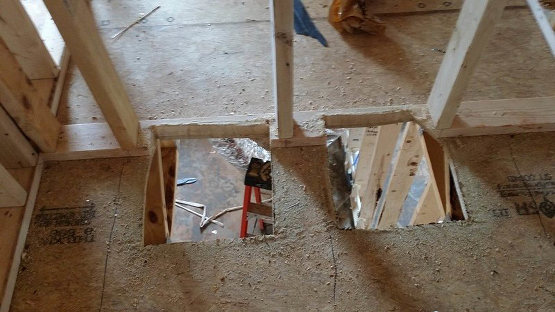 Excavated hole in subfloor surrounded by wooden beams, construction materials, and a red and black ladder inside unfinished home.