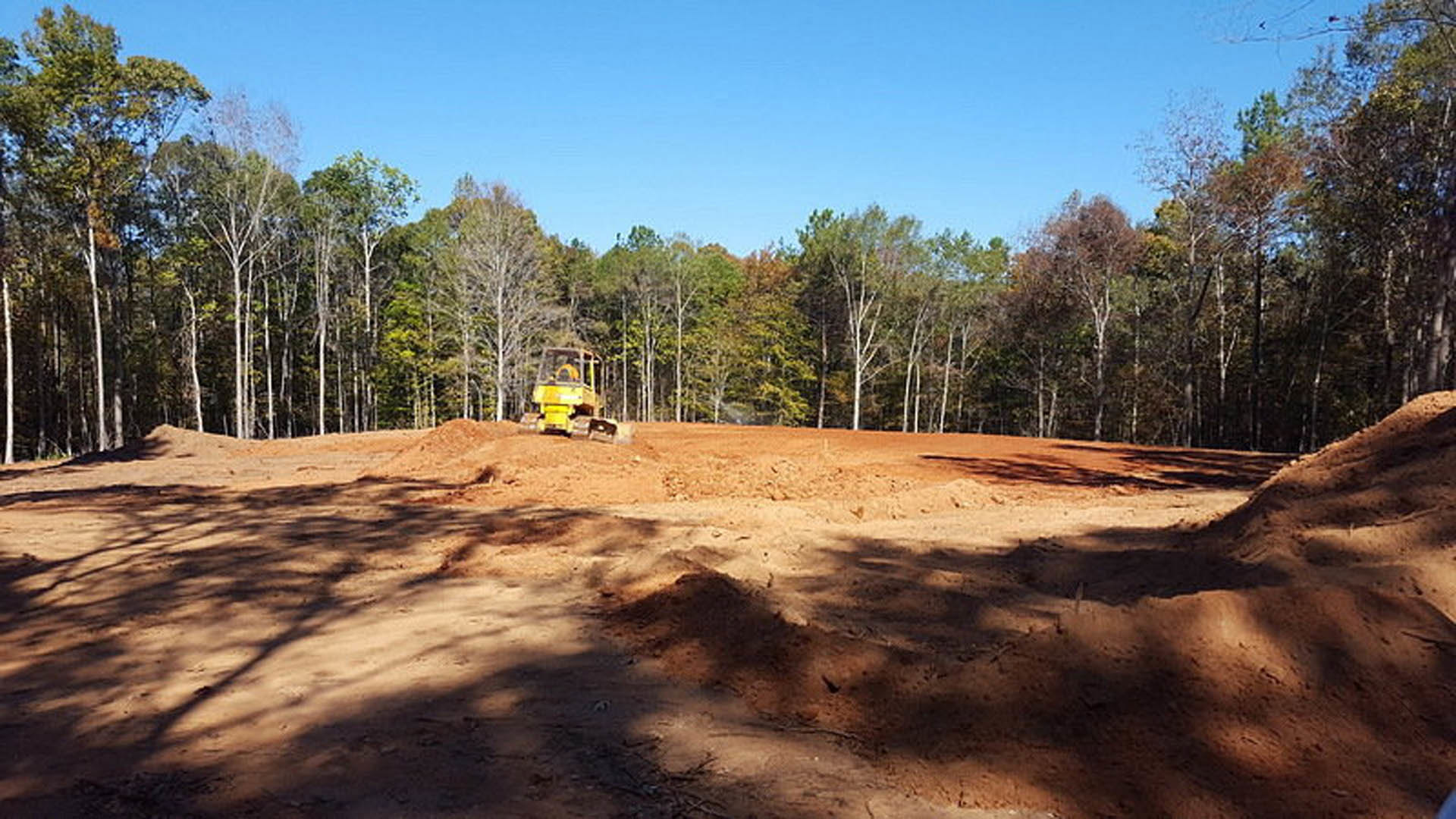 Yellow construction tractor parked on dirt lot with trees and blue sky in background