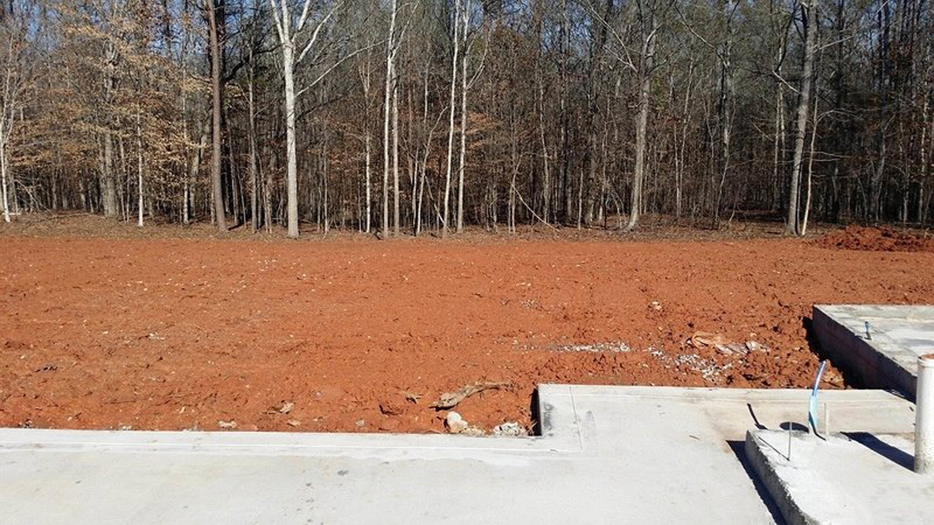 Dirt field bordered by trees, concrete slab with metal rod and flag, dirt road, concrete wall with pipe