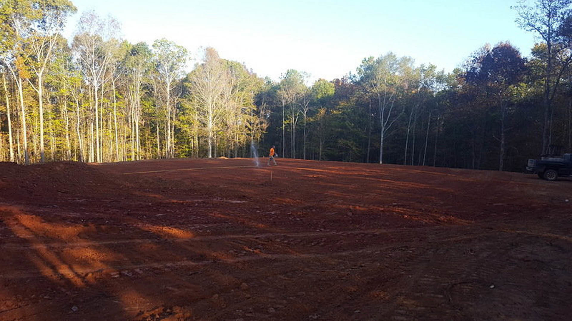 Person standing on a dirt field with scattered trees in the background under an open sky