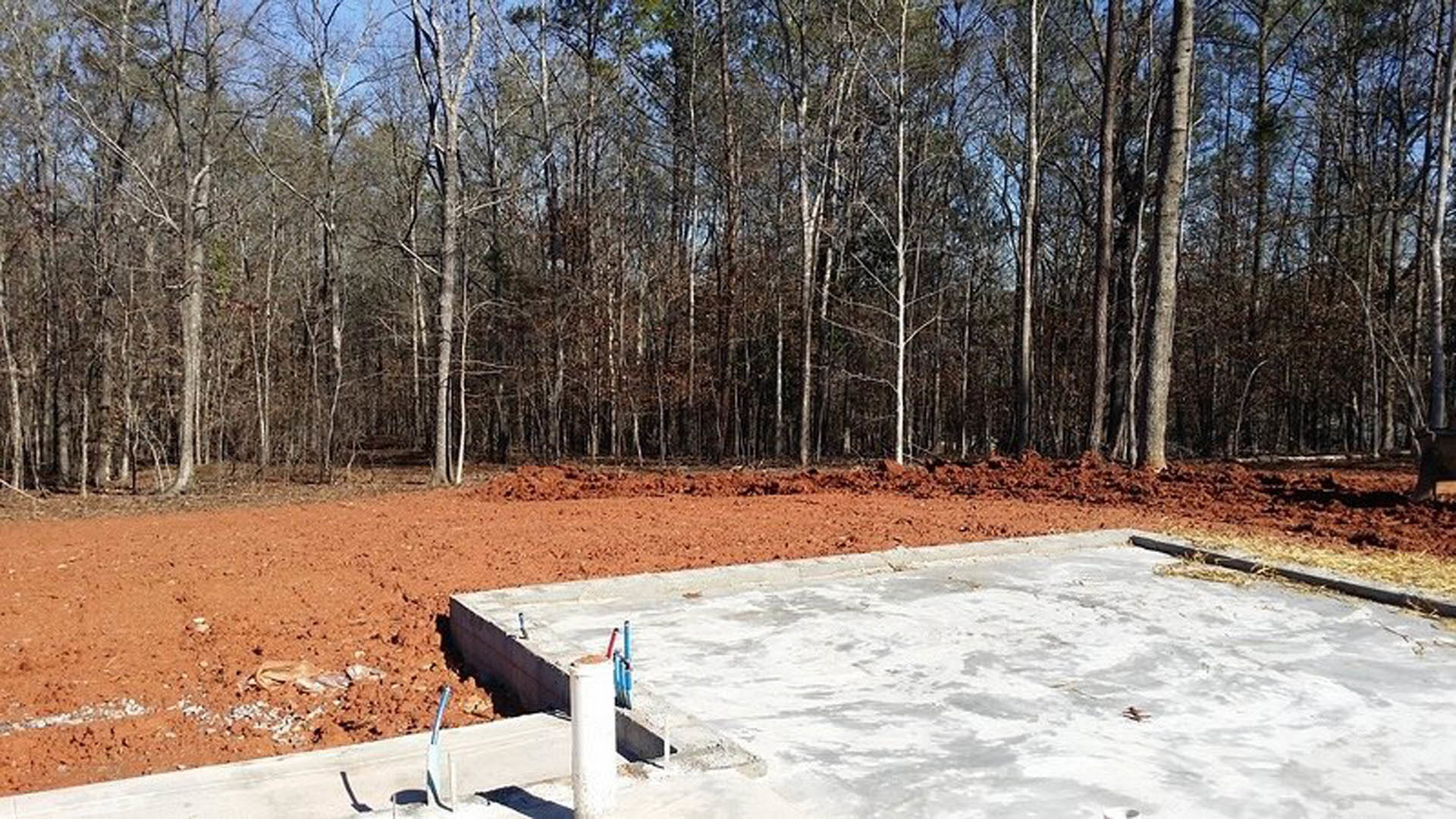 Concrete foundation surrounded by bare dirt, leafless trees, and patches of snow under a cloudy winter sky