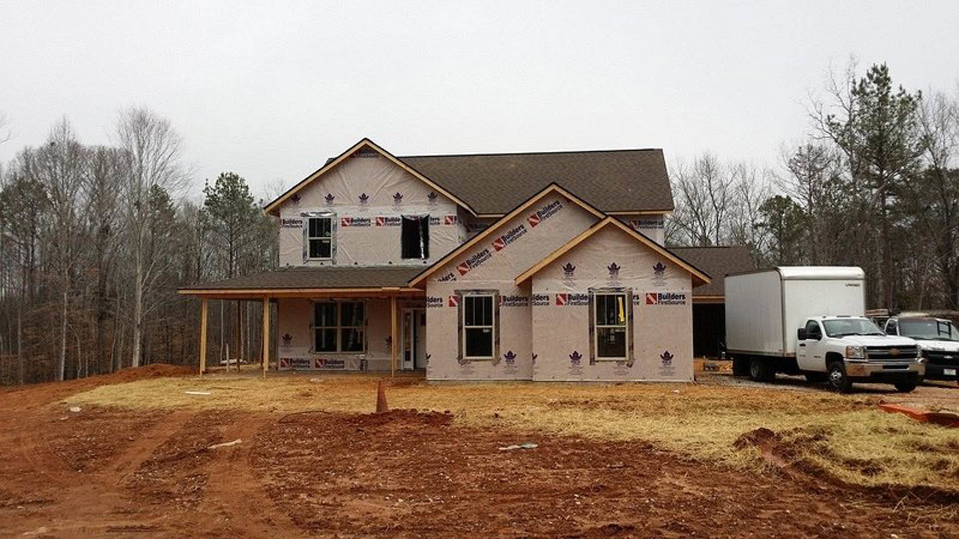 Framed house under construction with exposed wood, white truck parked on dirt driveway, surrounding trees and blue sky