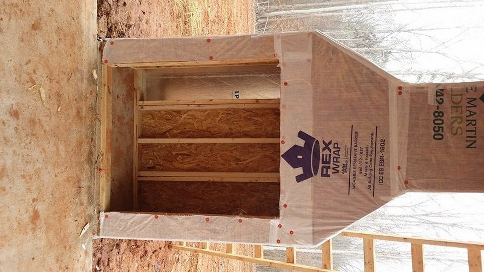 White plastic-wrapped wooden structure with exterior stairs, blue logo on tent covering, unfinished lumber walls, outdoor construction site