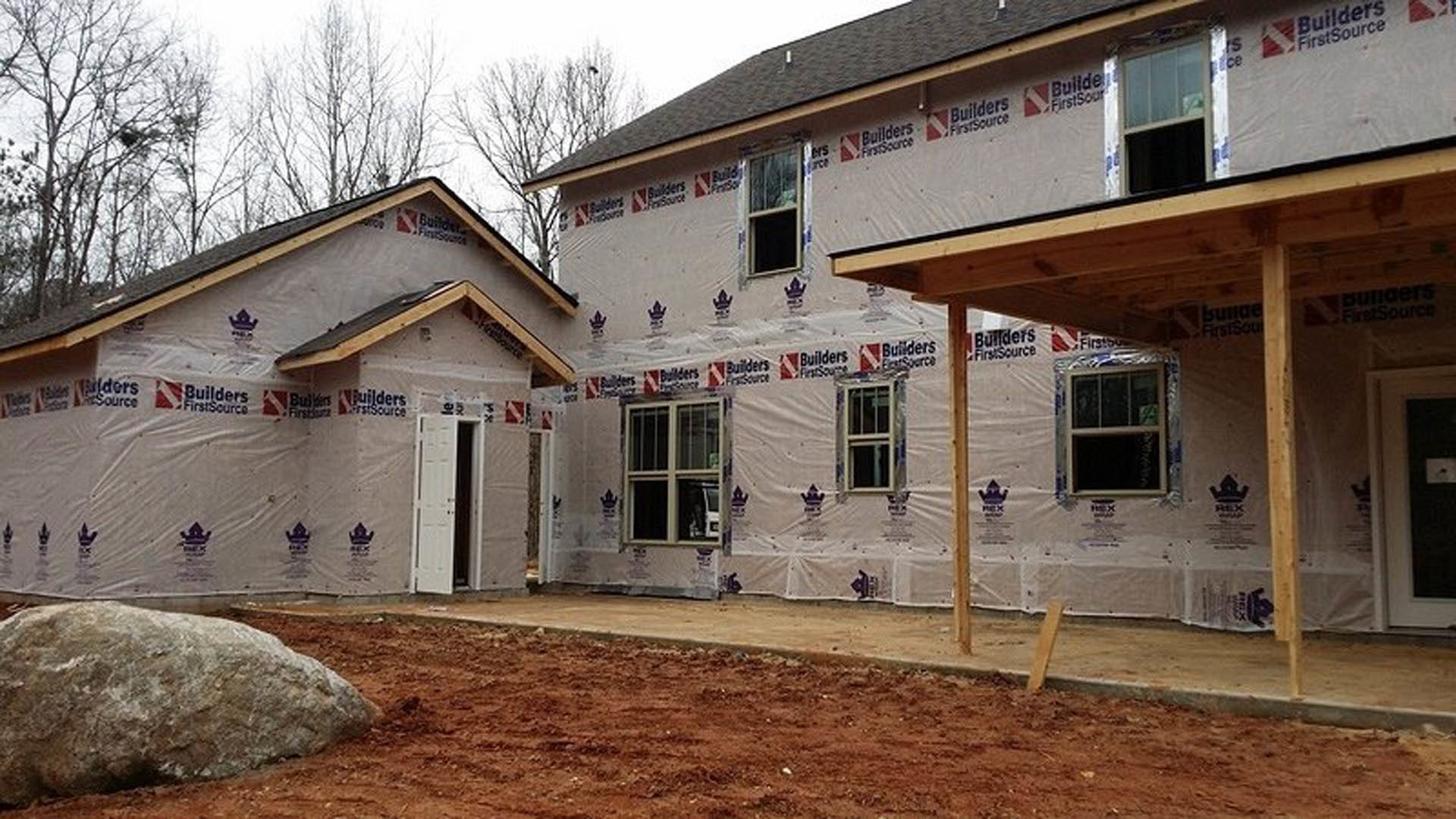 Partially built house with white framed windows, covered porch, exposed siding, large rock in dirt foreground, and construction materials scattered around