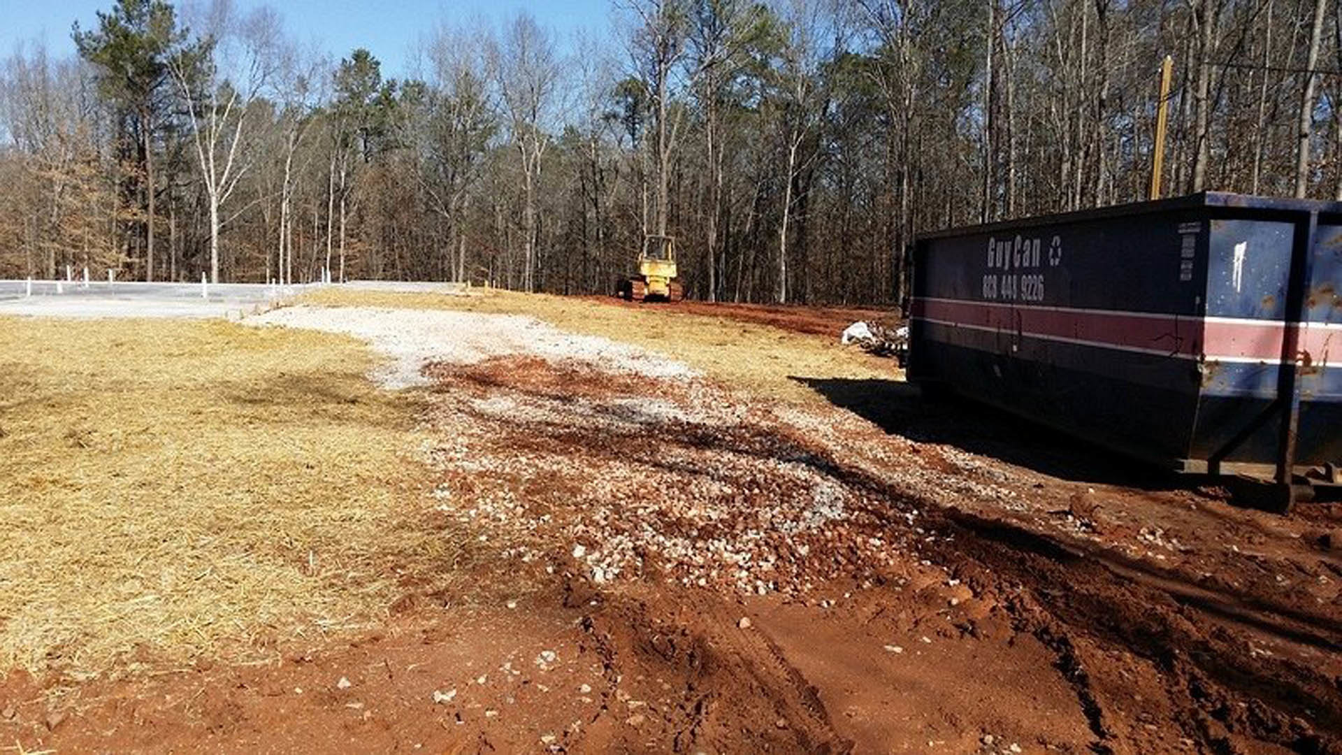 Yellow tractor parked on a dirt road beside grassy field, blue and red containers, and a yellow forklift near a wooden fence; trees and open sky in background.
