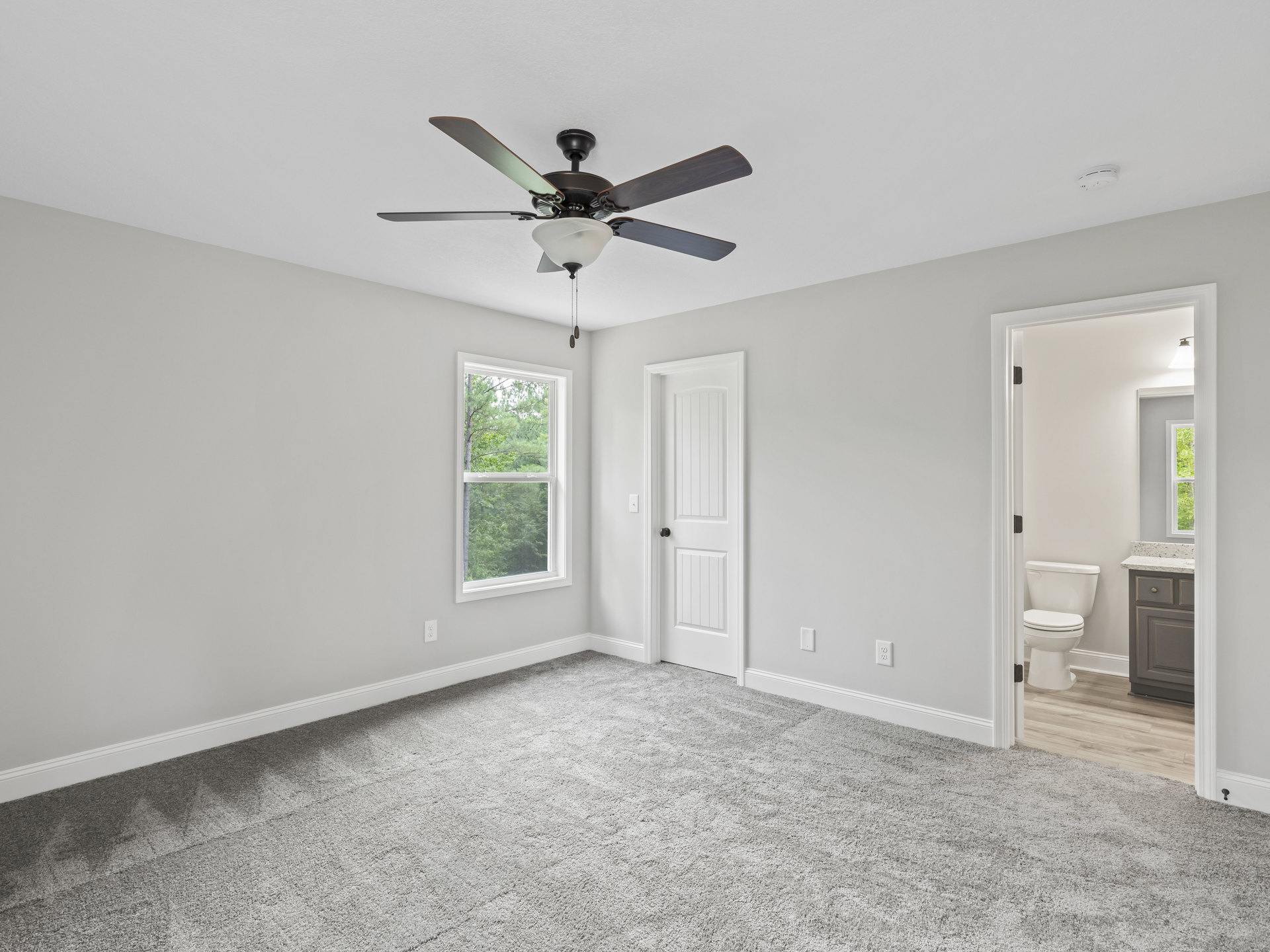 Carpeted room with white walls, ceiling fan with light fixture, white toilet with closed lid, window showing trees outside, white door with black handle