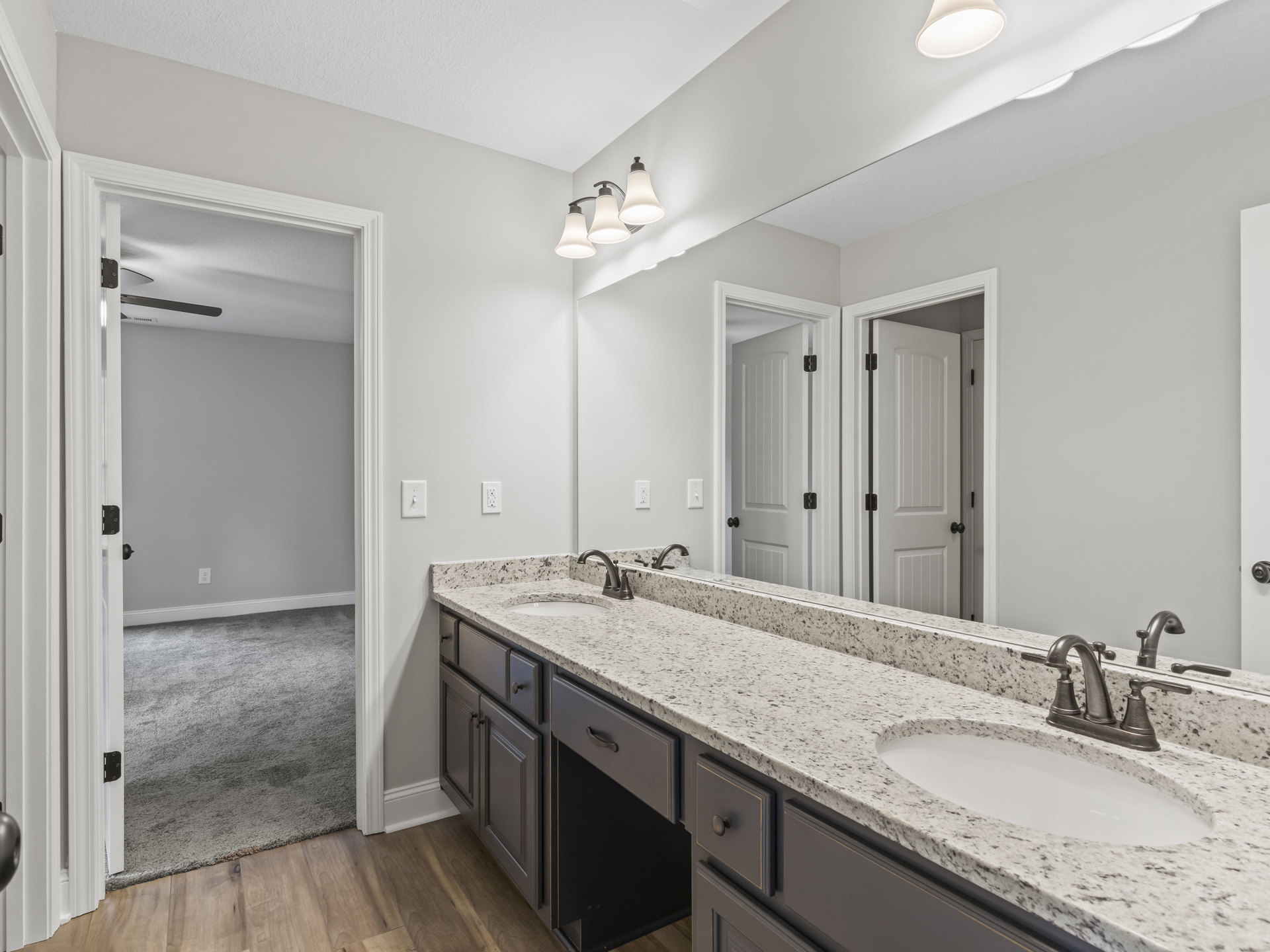 Bathroom with two undermount sinks set in a stone countertop, large framed mirror above, white cabinetry below, three-light fixture mounted over mirror, neutral tile backsplash