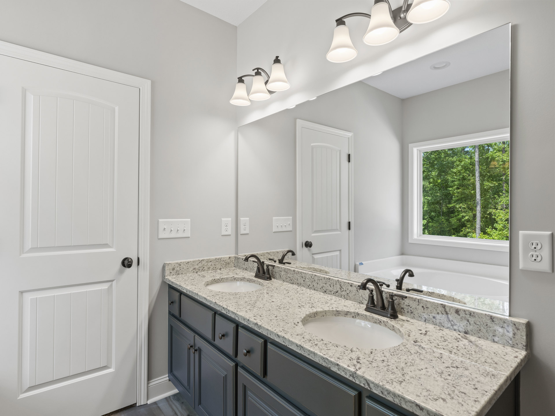 Bathroom with double vanity featuring black-speckled marble countertop, freestanding tub, white cabinetry, black hardware, large window overlooking trees, and white door with black