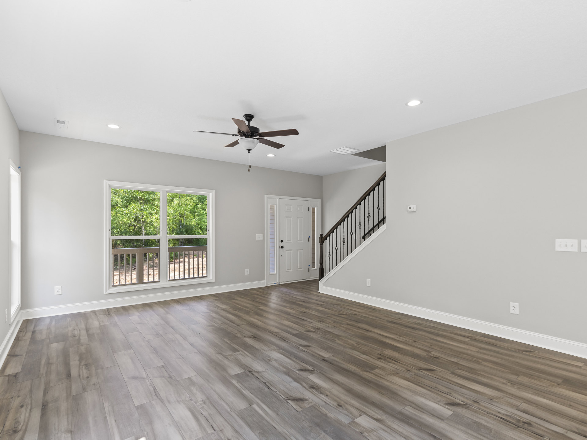 Ceiling fan with light fixture, hardwood floor, staircase with metal railings, white door with black handles, window showing trees outside, plaster walls and ceiling