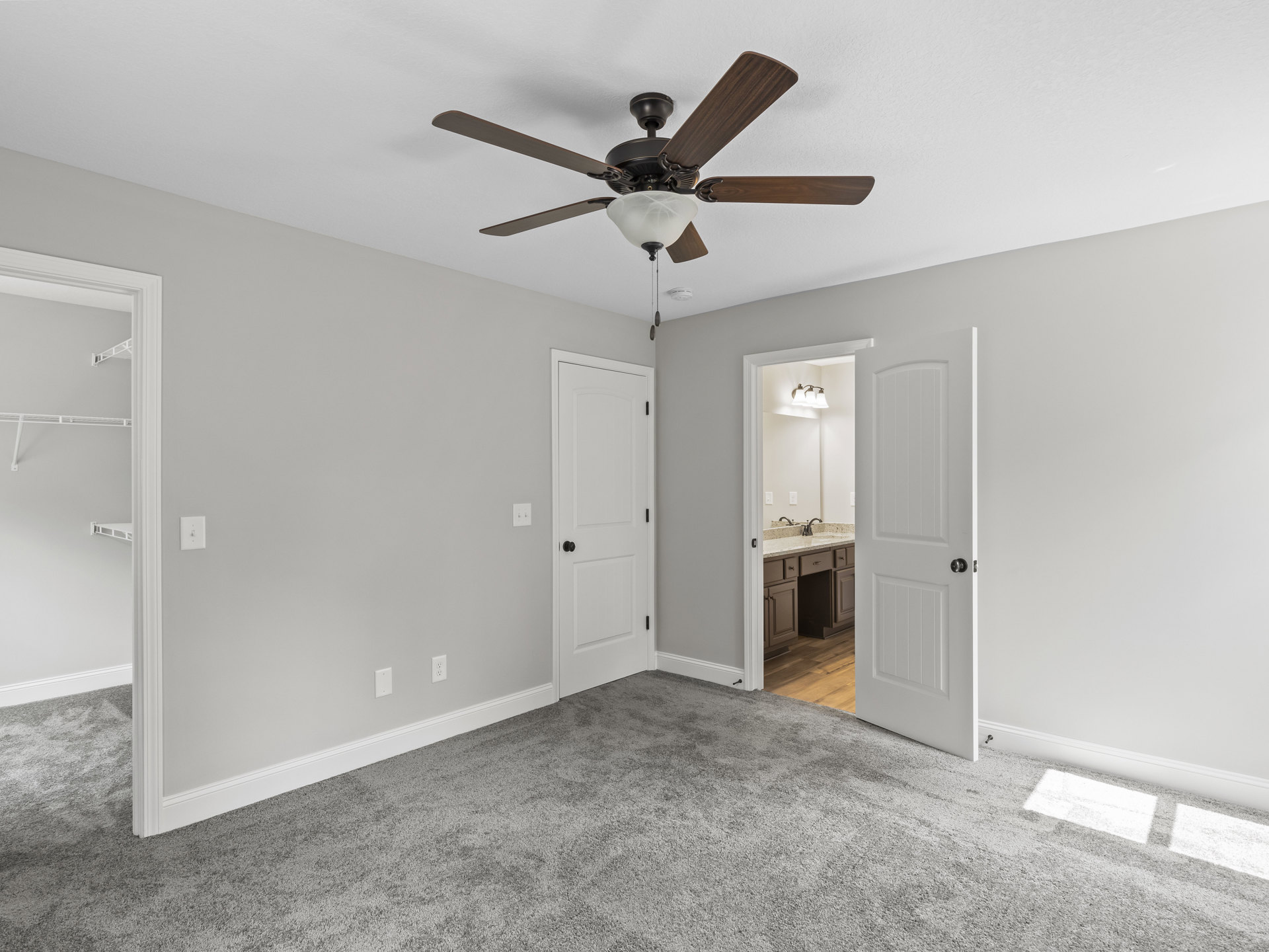 Bedroom with beige carpet, white walls, ceiling fan with light fixture, and adjacent bathroom visible through open white door with black hardware