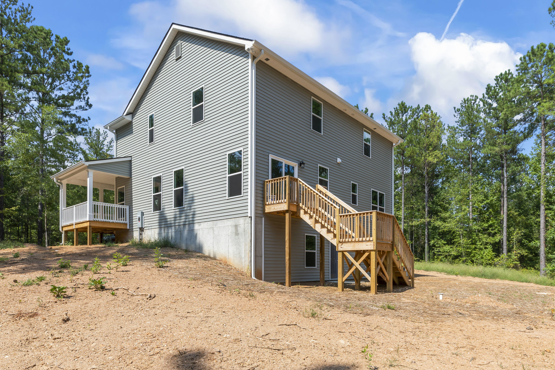 Two-story home with wood staircase leading to covered porch, large windows reflecting nearby tree, landscaped yard with plants and cloudy sky overhead