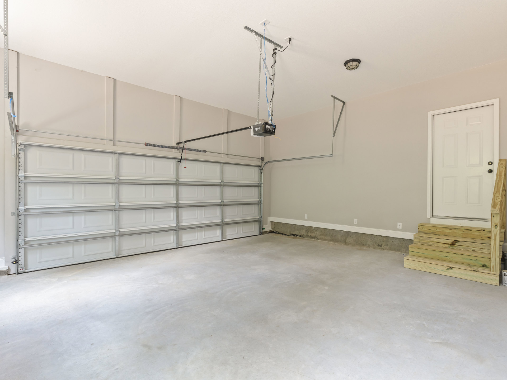 Garage interior with white plaster walls, wood staircase, ceiling-mounted light fixture, and lift garage door opener; visible wiring on metal stand near white door with illuminated