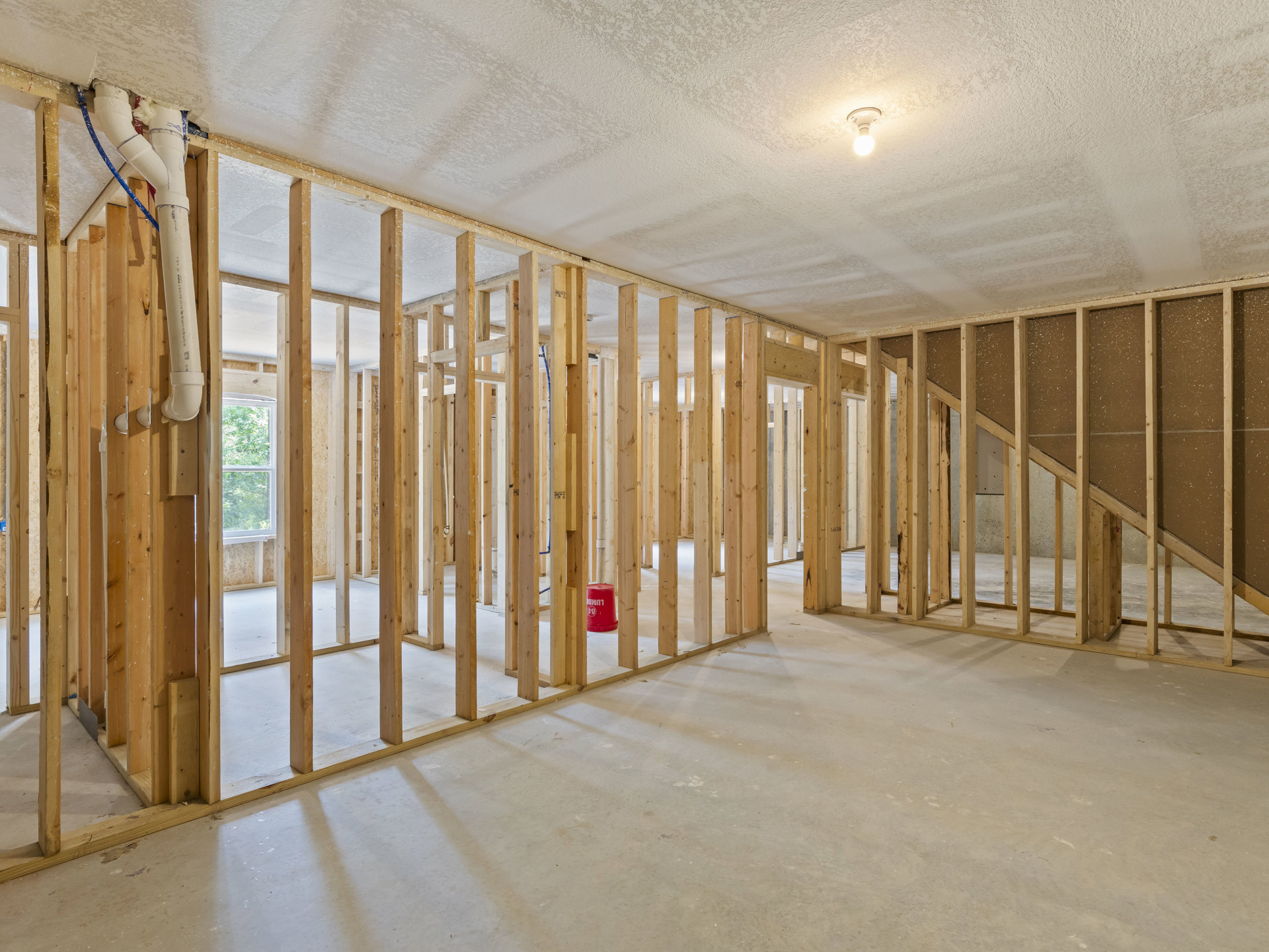 Wood-framed room under construction with exposed beams, white pipe labeled with blue and black text, single light bulb on ceiling, red bucket on unfinished floor, window showing