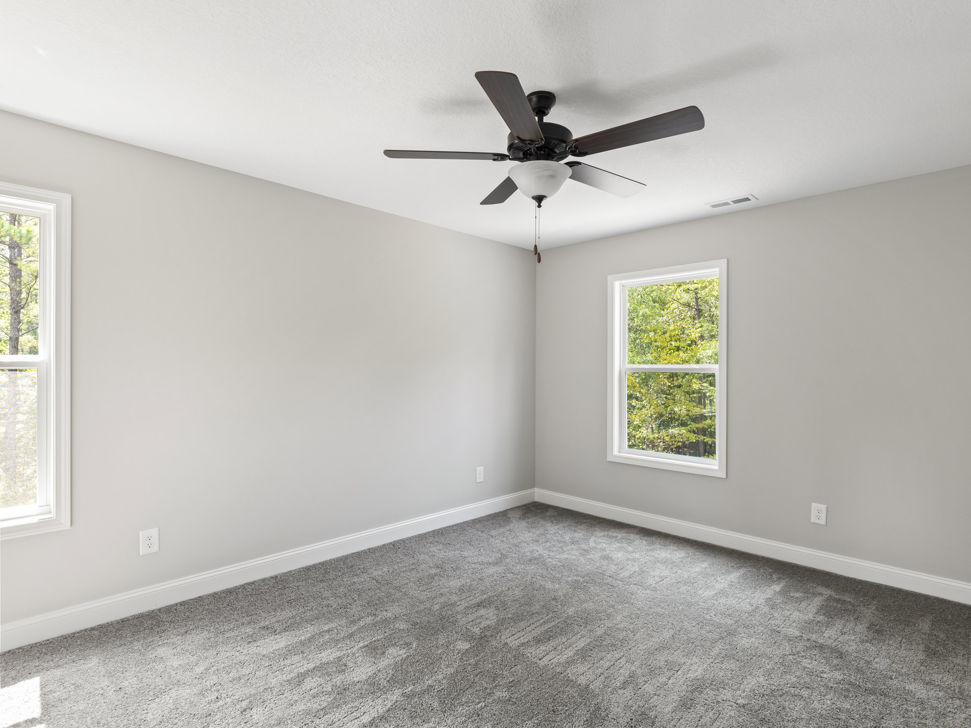 Ceiling fan with light fixture mounted above grey carpeted floor, large window revealing leafy trees outside, white plaster walls and ceiling