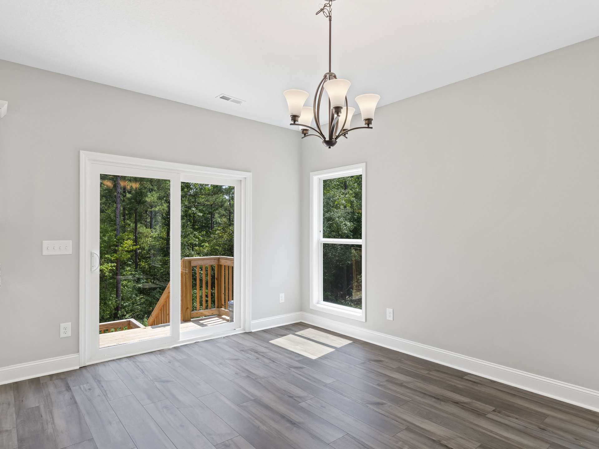 Chandelier with four lights hanging from a white ceiling, wooden flooring, white walls, large windows overlooking trees, wooden railing visible in background.