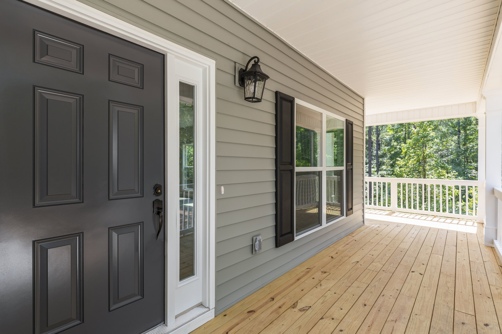 Wide wooden front porch with black door, adjacent window, and overhead light fixture; screen door opens onto natural wood decking.