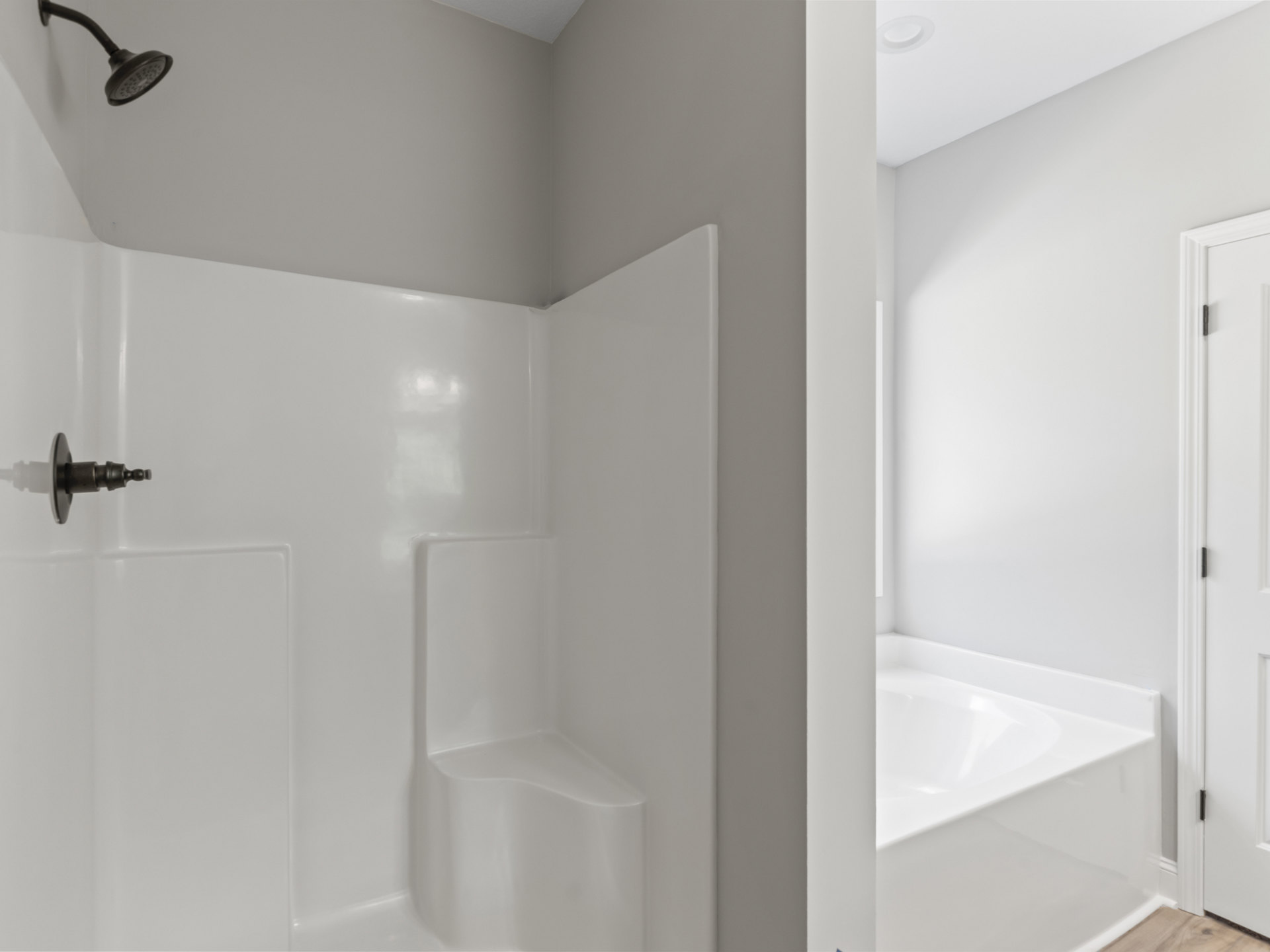 Freestanding white bathtub beside white tiled shower with black handle, metal shower head, white door with black hinge, and plaster walls