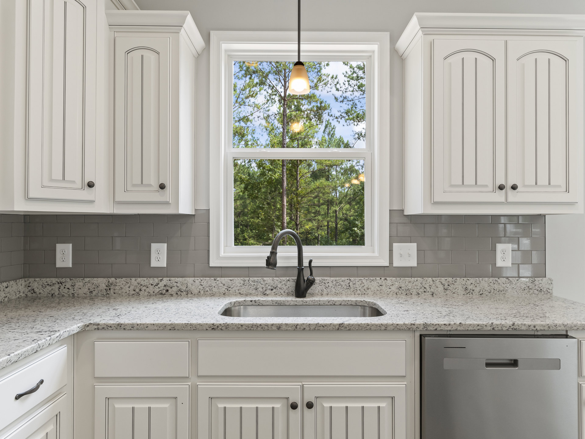 Modern kitchen featuring white cabinetry, quartz countertops, stainless steel dishwasher, curved chrome faucet, black vase on the counter, and a window overlooking leafy trees.