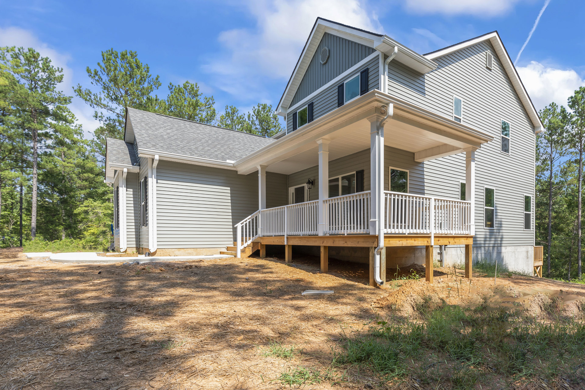 Two-story home with light siding, white porch railings, wood deck, large windows, and mature trees under blue sky
