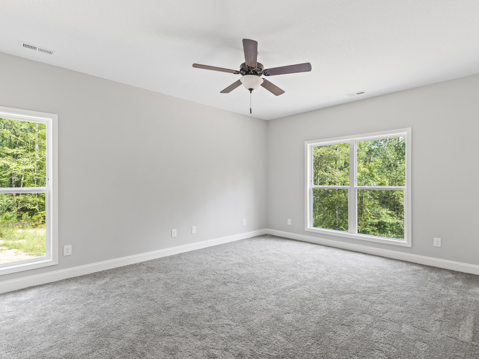 Neutral-toned carpeted room with white plaster walls, ceiling fan with light fixture, and large windows overlooking leafy trees