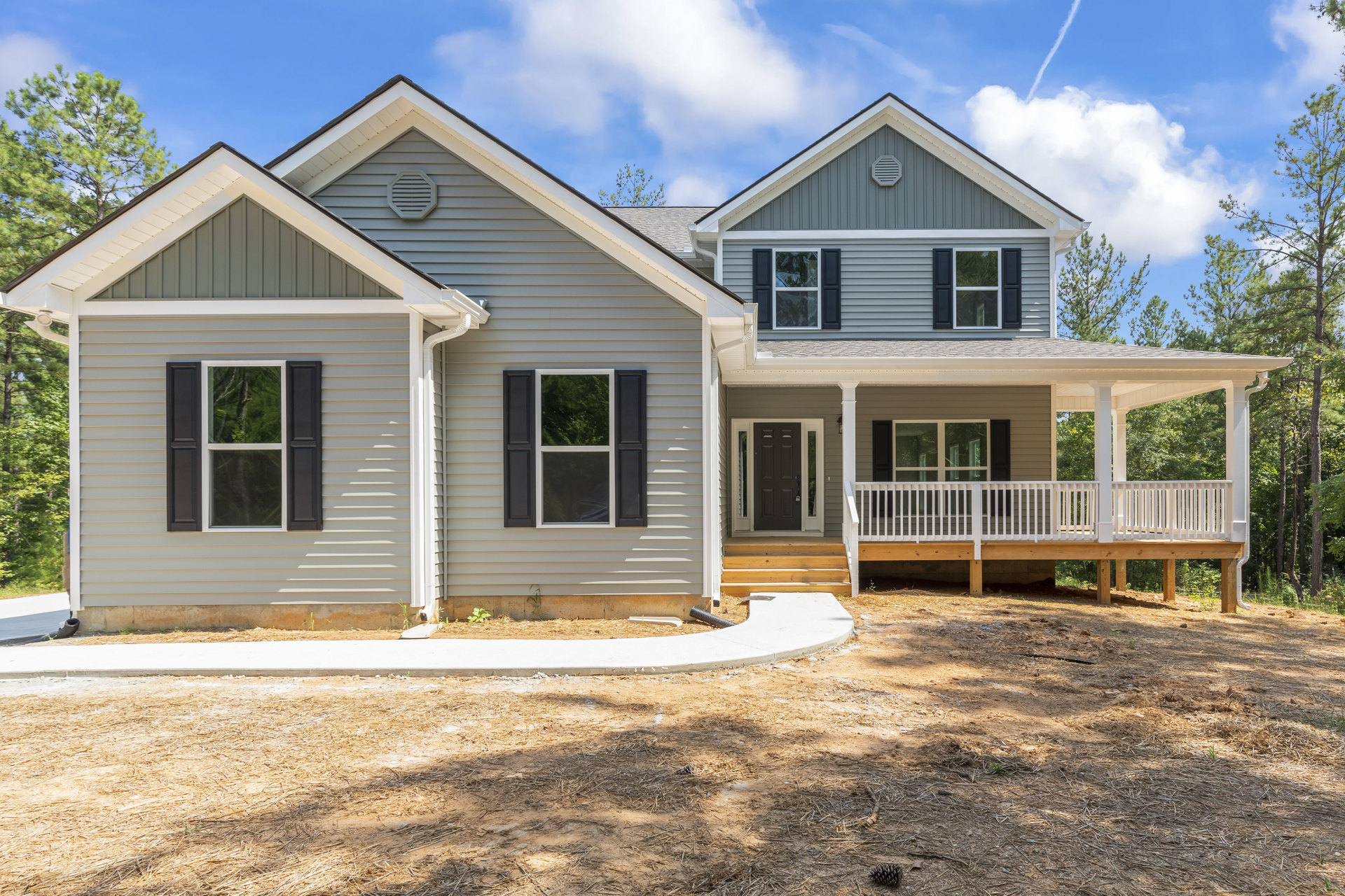 Two-story house with gray siding, white trim windows, covered front porch with white railings, concrete driveway, and walkway bordered by landscaping.