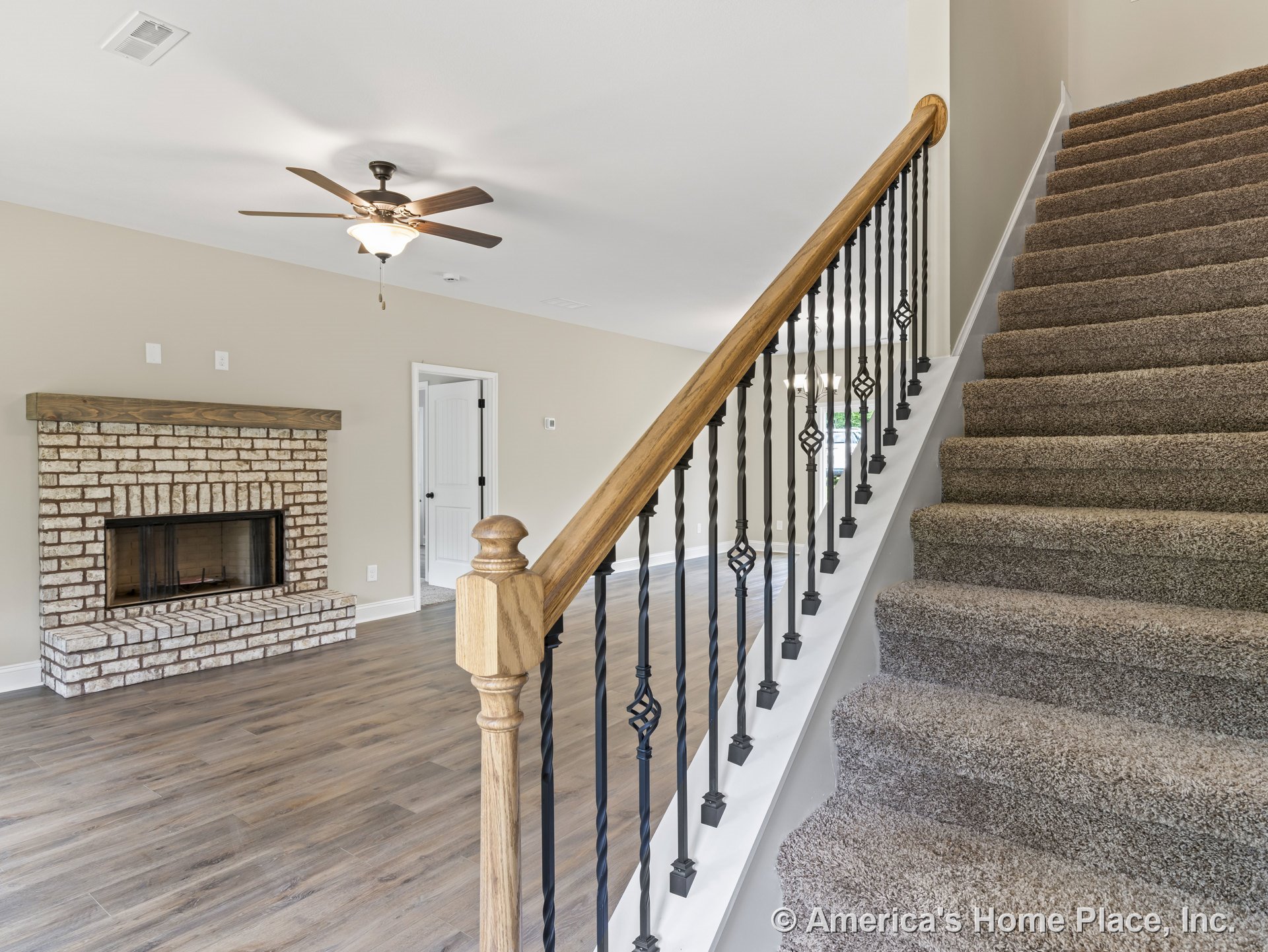 Brick fireplace with wood mantel, carpeted staircase with iron balusters, wood-look plank flooring, ceiling fan with light, and neutral wall paint in living room.