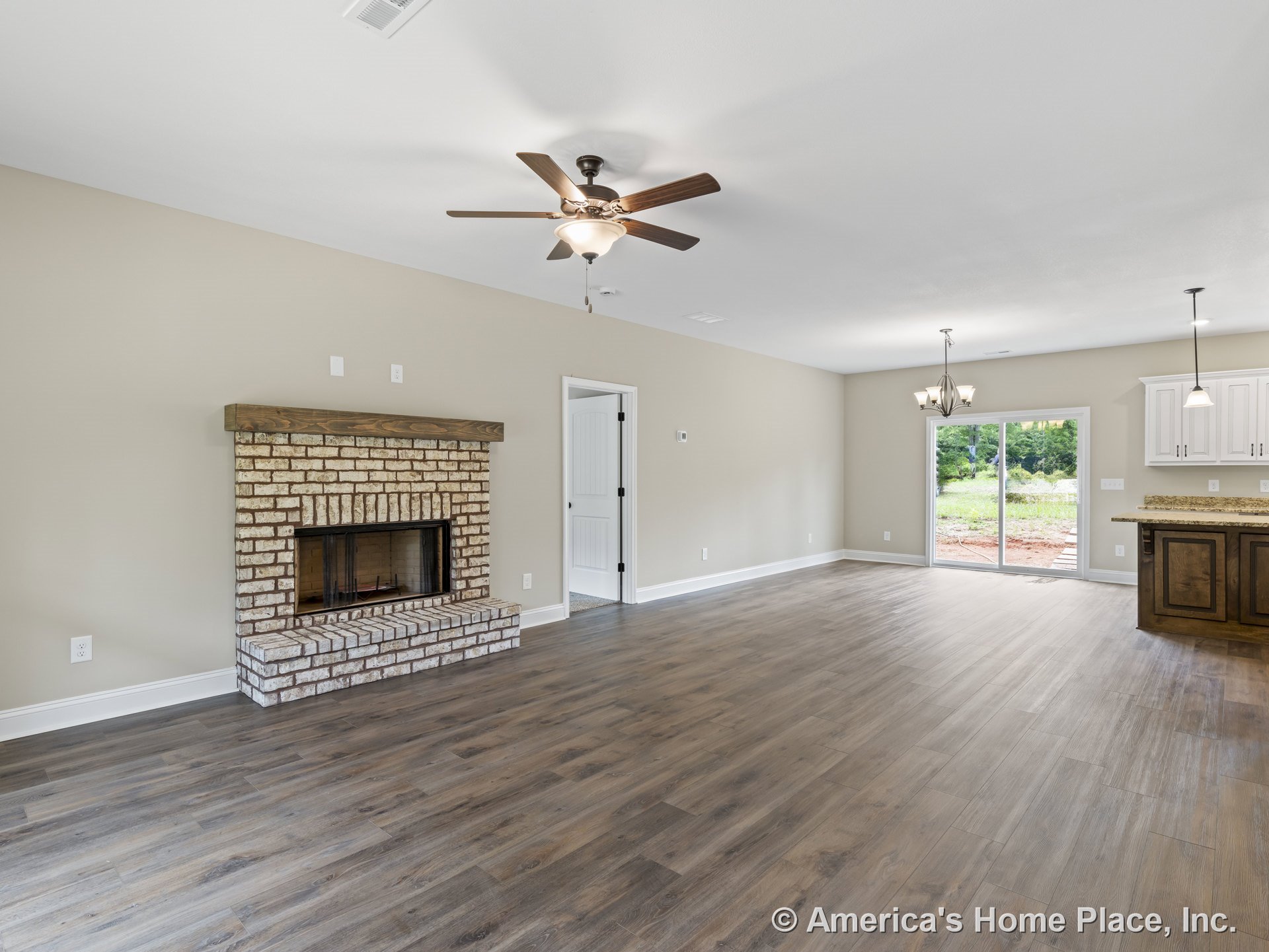 Brick fireplace with wood mantel, wood-look plank flooring, open layout connecting living area to kitchen with white cabinetry and granite countertops, ceiling fan with light