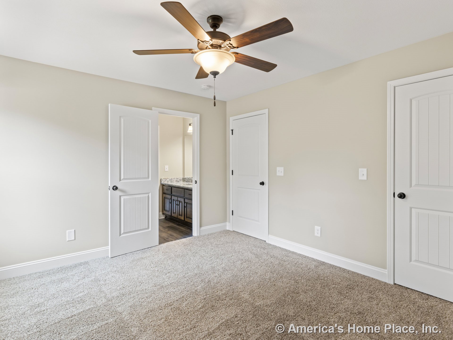 Beige carpeted bedroom with paneled white doors, ceiling fan and light fixture, neutral wall color, white baseboard trim, and bathroom vanity visible through open doorway.