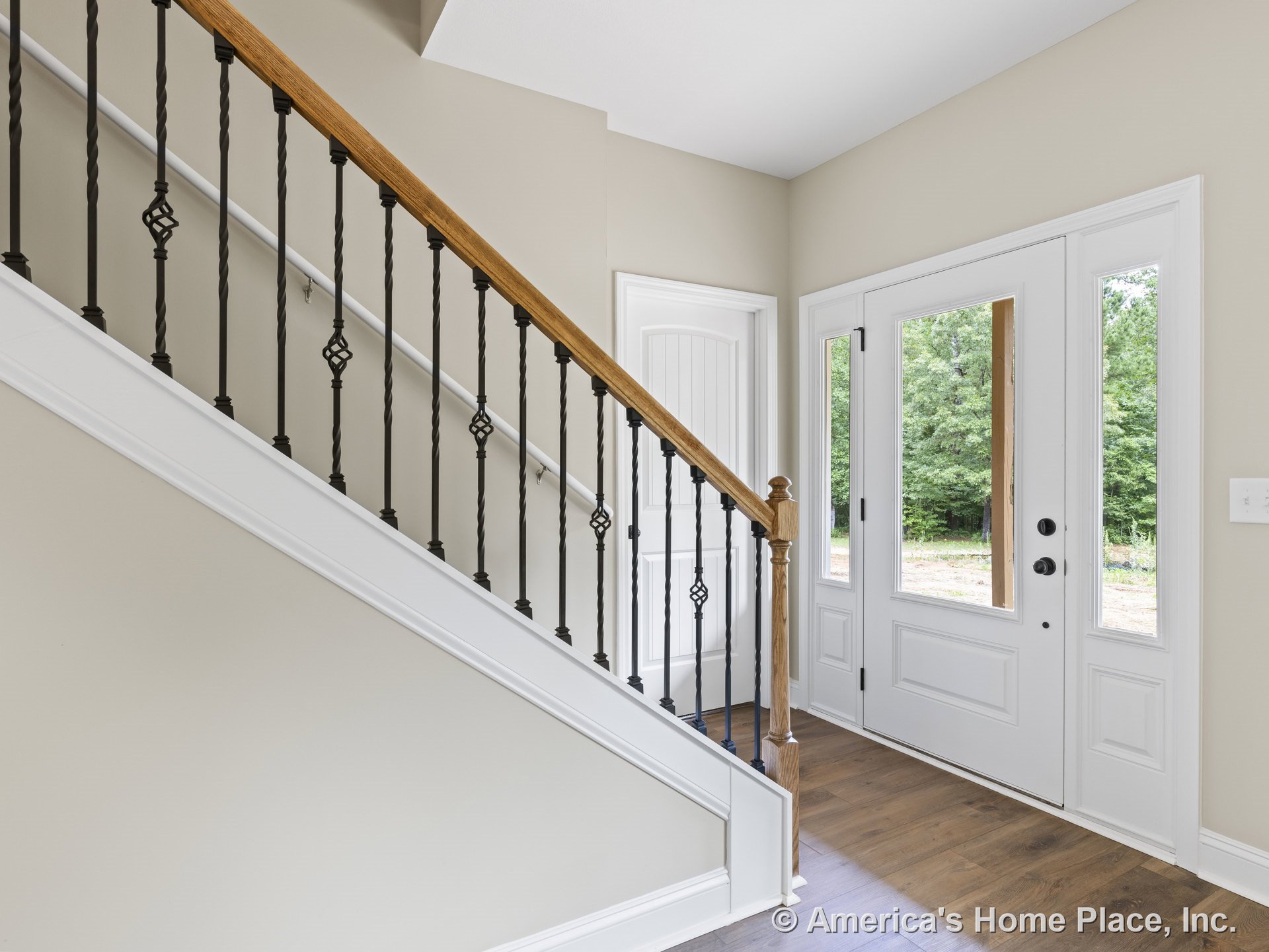 White front door with glass panel sidelights, hardwood flooring, staircase featuring wrought iron balusters and wood handrail, neutral wall paint, and white interior trim.